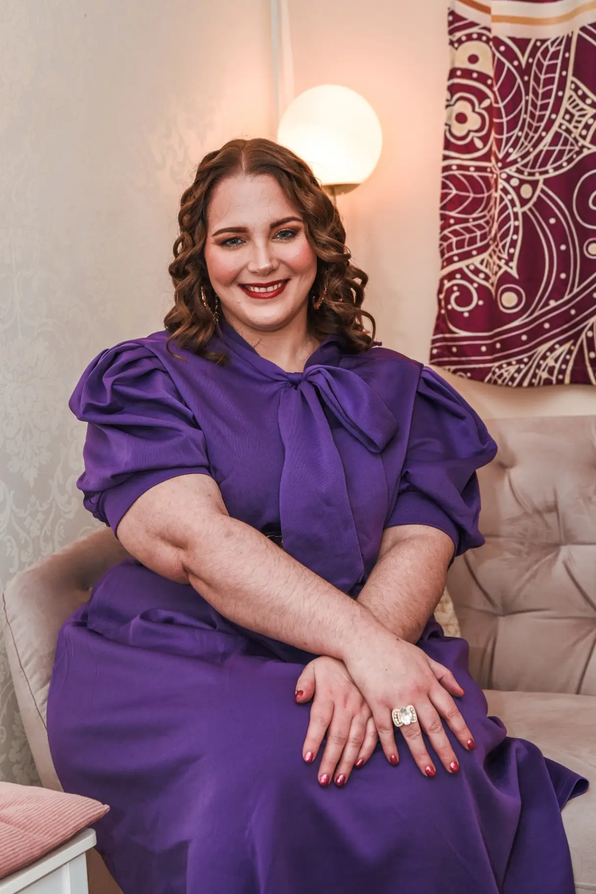 A woman with curly hair smiles warmly, wearing a purple dress with puffed sleeves and a large bow. Her hands are gently clasped in front